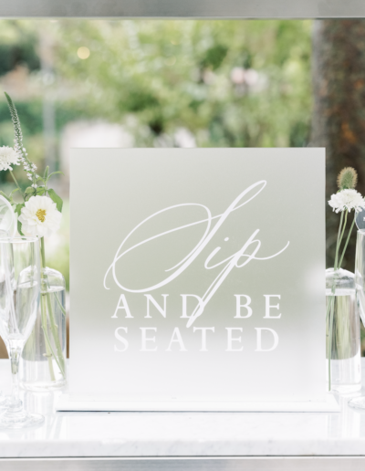 Decorative sign with the text "Sip and Be Seated" displayed on a table with flowers and glassware.